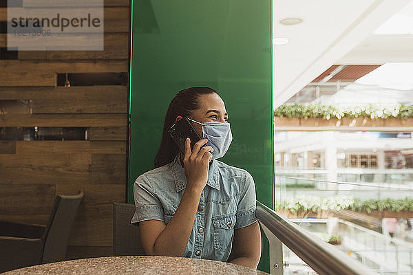 Junge Frau mit Maske  die in einem Einkaufszentrum über ein Smartphone spricht