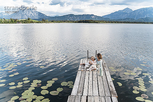 Mittlere erwachsene Frau und Tochter sitzen auf dem Steg über dem See gegen den Himmel