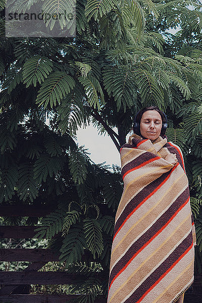 Frau mit geschlossenen Augen  eingewickelt in eine Decke  hört Musik über Kopfhörer am Akazienbaum (Acacia dealbata)
