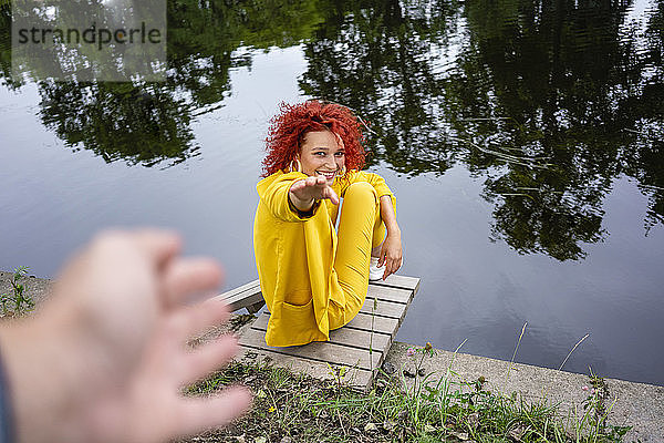 Junge Frau mit lockigem Haar und gelbem Anzug sitzt am Flussufer und greift nach der Hand eines Mannes.