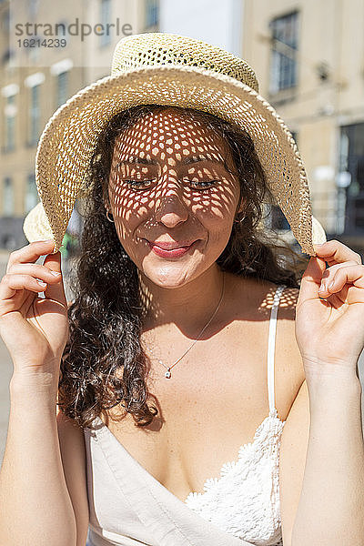 Nahaufnahme einer lächelnden schönen Frau mit geschlossenen Augen  die einen Hut in der Stadt an einem sonnigen Tag trägt
