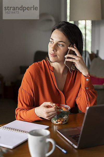 Geschäftsfrau  die mit ihrem Handy telefoniert  während sie zu Hause Salat isst