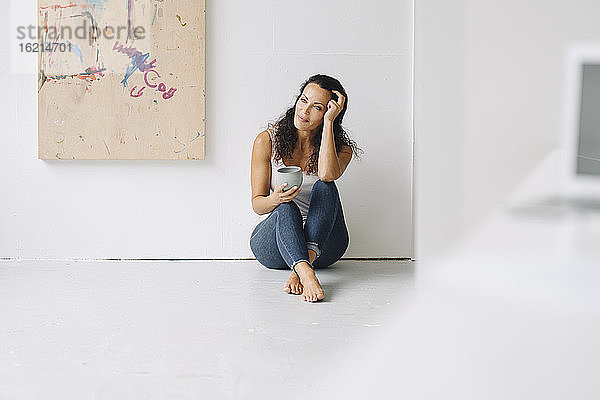 Nachdenkliche Frau mit Kaffeetasse auf dem Boden sitzend gegen die Wand in einem Loft