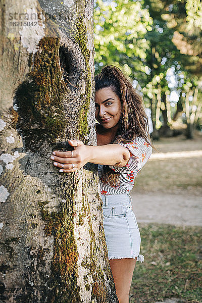 Lächelnde junge Frau  die einen Baumstamm hält  während sie im Park steht