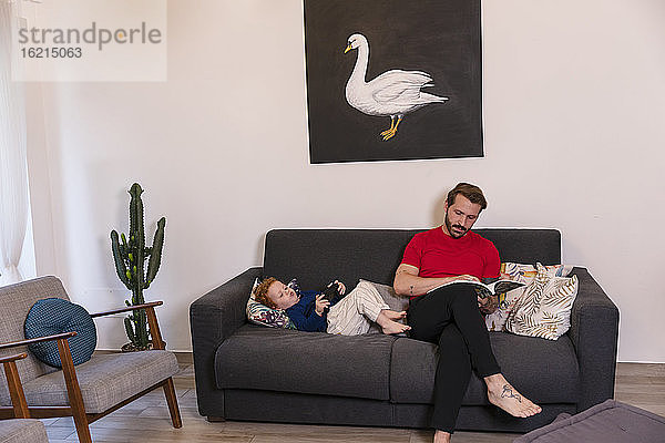 Mann liest eine Zeitschrift  während sein Sohn zu Hause auf dem Sofa mit einem Joystick spielt