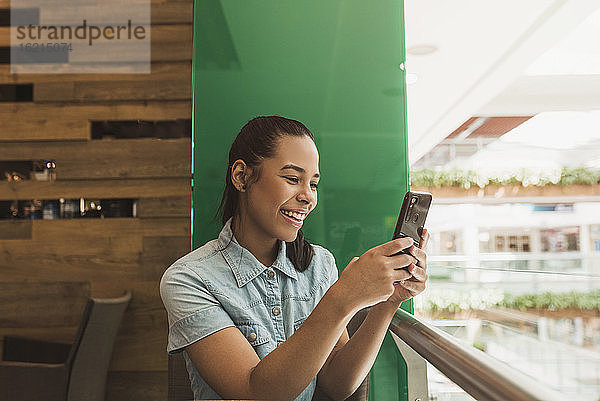 Fröhliche junge Frau  die ihr Smartphone benutzt  während sie an einem Geländer in einem Einkaufszentrum sitzt