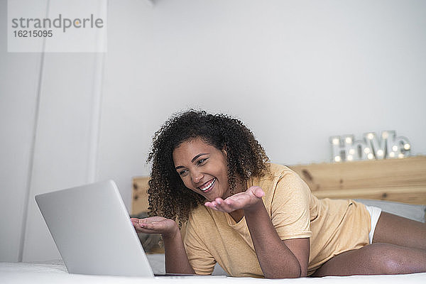 Fröhliche junge Frau bei einer Videokonferenz über einen Laptop  während sie zu Hause auf dem Bett liegt