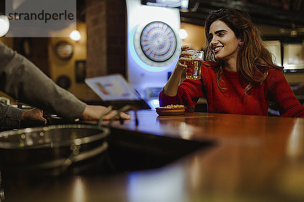 Lächelnde schöne Frau hält Bierglas im Gespräch mit Barkeeper im Restaurant