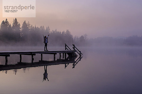 Frau steht mit erhobenen Armen auf einem Steg am Seeufer in der nebligen Morgendämmerung