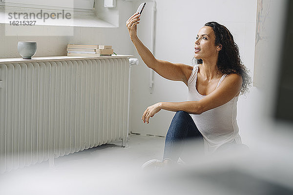Mittlere erwachsene Frau  die ein Selfie mit ihrem Smartphone macht  während sie zu Hause auf dem Dachboden sitzt
