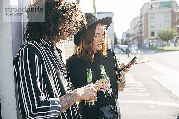 Paar  das Bierflaschen hält und ein Smartphone benutzt  während es auf der Straße in der Stadt steht