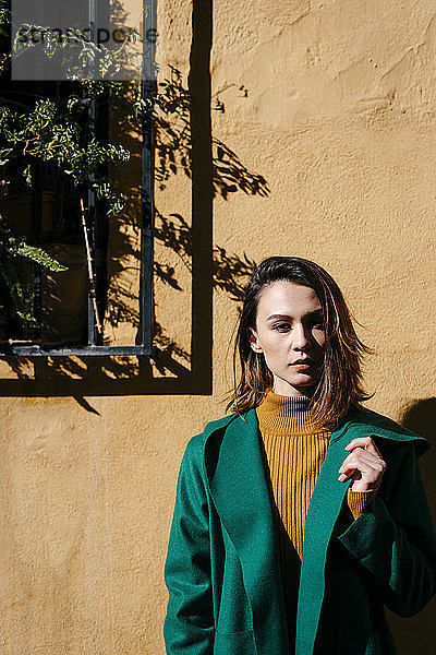 Schöne Frau in grüner Jacke steht an einer Wand an einem sonnigen Tag