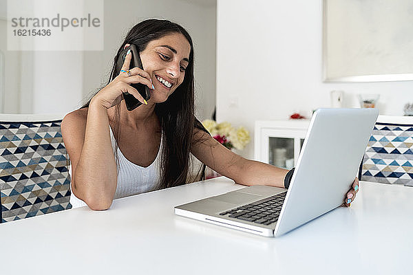 Lächelnde Frau im mittleren Erwachsenenalter  die über ein Mobiltelefon spricht  während sie einen Laptop auf einem Tisch zu Hause benutzt