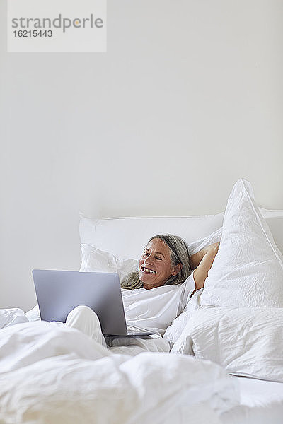 Lächelnde Frau  die einen Laptop benutzt  während sie zu Hause auf dem Bett liegt