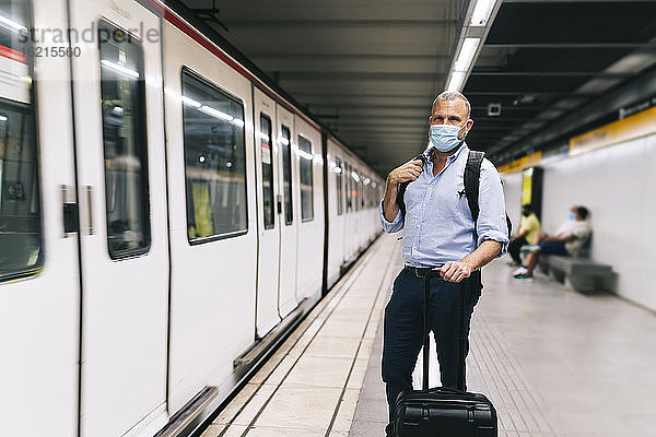 Geschäftsmann mit Gesichtsmaske  während er mit Gepäck auf dem Bahnsteig der U-Bahn steht