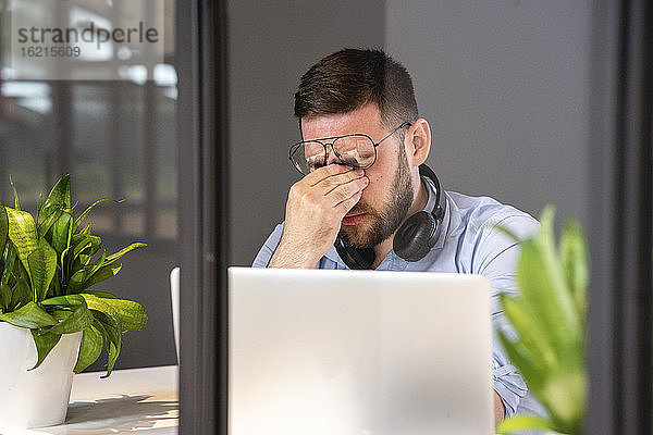 Müde gutaussehende männliche Fachkraft mit Laptop durch Glaswand in kreativem Coworking-Büro gesehen