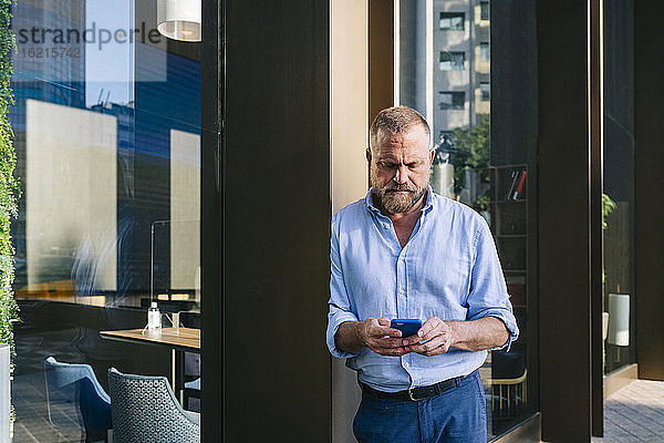 Geschäftsmann  der sein Smartphone benutzt und sich an die Wand eines Bürogebäudes lehnt