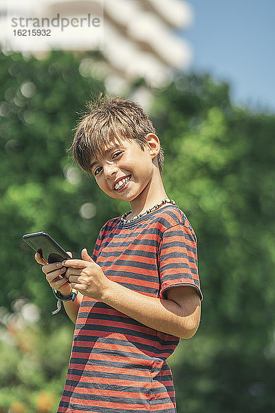 Lächelnder Junge  der sein Smartphone benutzt  während er an einem sonnigen Tag vor einem Baum steht