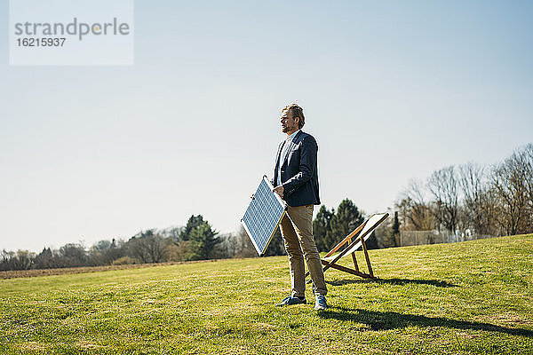 Geschäftsmann  der wegschaut  während er ein Solarmodul mit einem Stuhl auf dem Rasen eines Parks gegen den klaren Himmel an einem sonnigen Tag hält