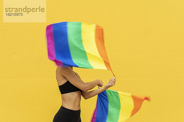 Frau schwenkt Regenbogenfahnen und steht vor einer gelben Wand in der Stadt