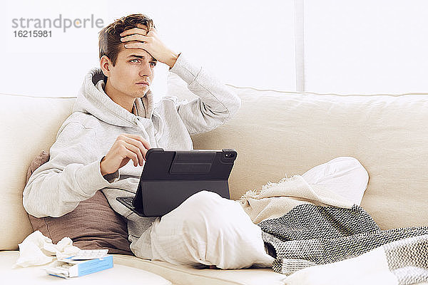 Junger Mann mit Kopf in den Händen  der auf dem Sofa zu Hause ein digitales Tablet benutzt