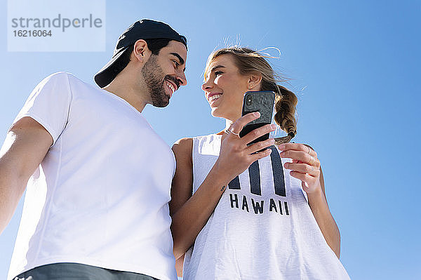 Lächelnde Frau  die ihr Smartphone hält und ihren Freund gegen den klaren blauen Himmel ansieht