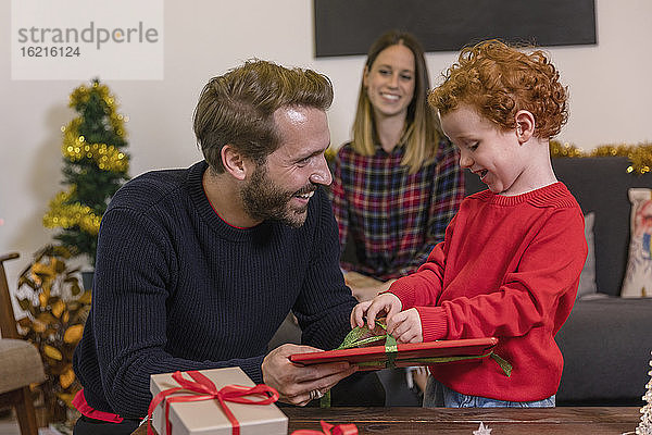 Lächelnde Frau betrachtet den Vater  der seinem Sohn im Wohnzimmer ein Weihnachtsgeschenk macht