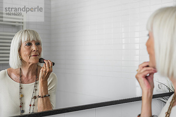 Ältere Frau trägt Make-up auf  während sie zu Hause in den Spiegel schaut