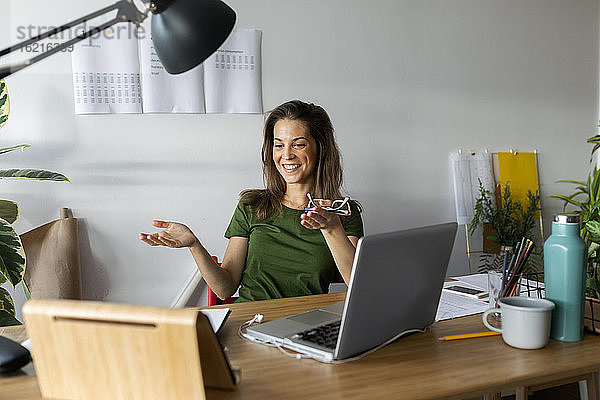 Lächelnde junge Frau gestikuliert  während sie auf ein digitales Tablet auf dem Schreibtisch im Heimbüro schaut