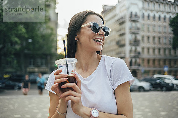 Fröhliche schöne Frau mit Sonnenbrille  die ein Erfrischungsgetränk hält  während sie in der Stadt steht