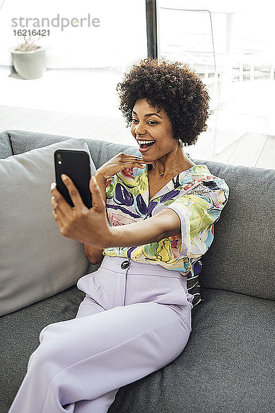 Aufgeregte Frau  die ein Selfie mit ihrem Smartphone macht  während sie auf dem Sofa im Penthouse sitzt