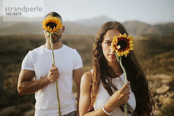 Frau und Mann verstecken ihr Gesicht mit Sonnenblumen bei Sonnenuntergang