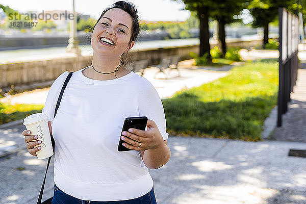 Porträt einer glücklichen  kurvigen jungen Frau mit Getränk zum Mitnehmen und Mobiltelefon in der Stadt