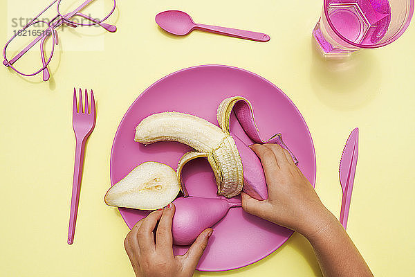 Hand eines kleinen Mädchens  das eine pinkfarbene Birne und eine Banane von einem Plastikteller aufhebt