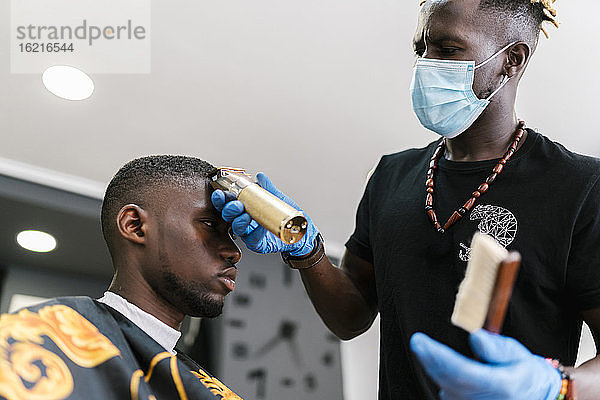 Männlicher Friseur mit Maske schneidet einem Kunden die Haare mit einem Rasiermesser im Friseursalon