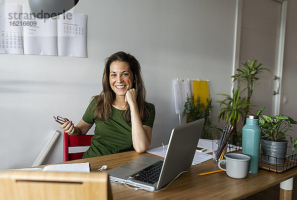 Lächelnde Geschäftsfrau sitzt an einem Schreibtisch an der Wand im Büro zu Hause