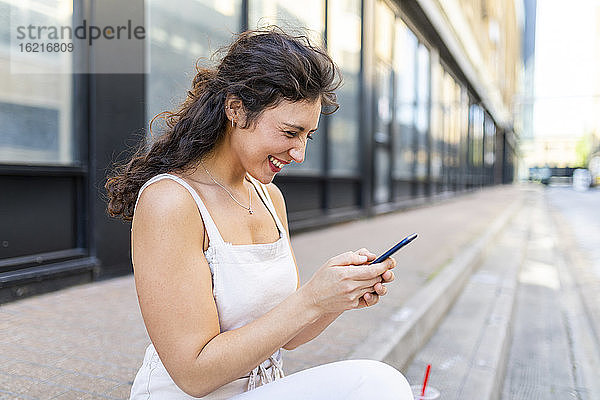 Fröhliche junge Frau benutzt ihr Smartphone  während sie auf einer Treppe in der Stadt sitzt