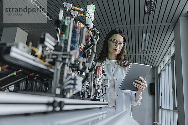 Junge Frau benutzt ein digitales Tablet  während sie an einer Maschine im Labor steht