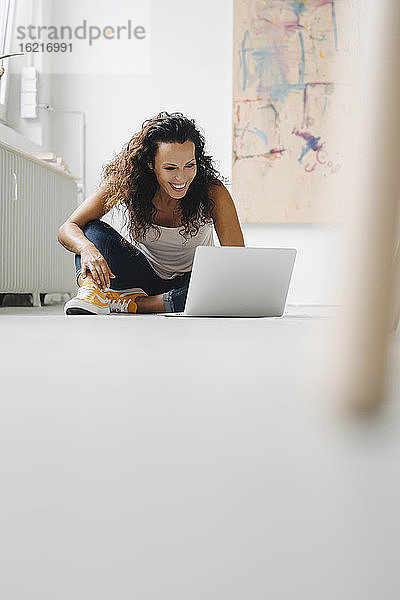 Fröhliche Frau im mittleren Erwachsenenalter  die einen Laptop benutzt  während sie zu Hause auf dem Boden sitzt