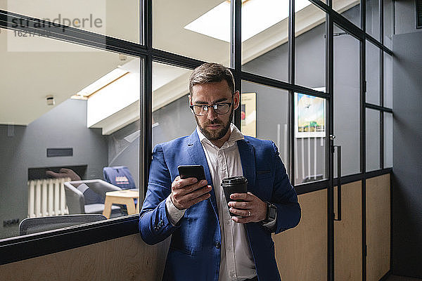 Geschäftsmann hält Einweg-Kaffeebecher  während er sein Smartphone an der Glaswand eines Coworking-Büros benutzt