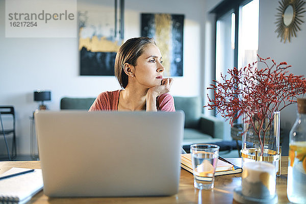Nachdenkliche junge Frau mit Laptop auf dem Schreibtisch  die wegschaut  während sie zu Hause sitzt