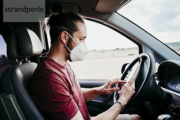 Mann benutzt Smartphone  während er im Auto eine Gesichtsschutzmaske trägt