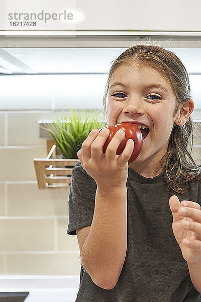 Nahaufnahme eines glücklichen Mädchens  das eine Tomate isst  während es zu Hause in der Küche sitzt