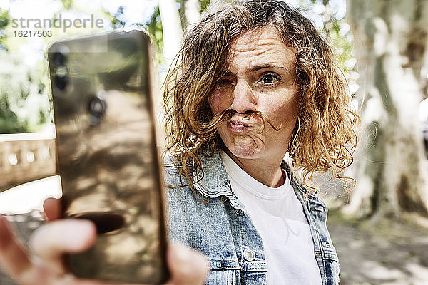 Nahaufnahme einer Frau  die ein Gesicht macht  während sie ein Selfie mit ihrem Smartphone im Park macht