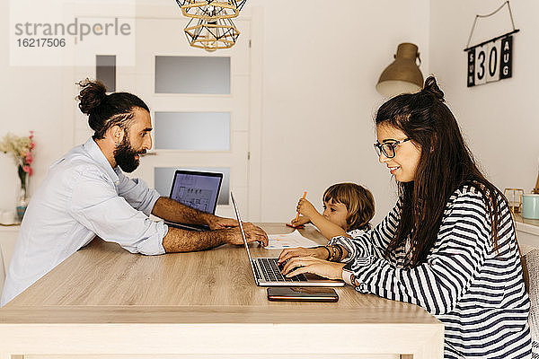 Frau arbeitet am Laptop  während Vater und Tochter zu Hause am Esstisch malen