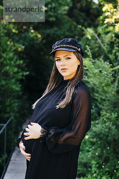 Schöne schwangere Frau steht mit Händen auf dem Bauch im Park