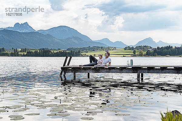 Vater und Tochter betrachten die Aussicht  während sie sich auf dem Steg über dem See vor den Bergen entspannen