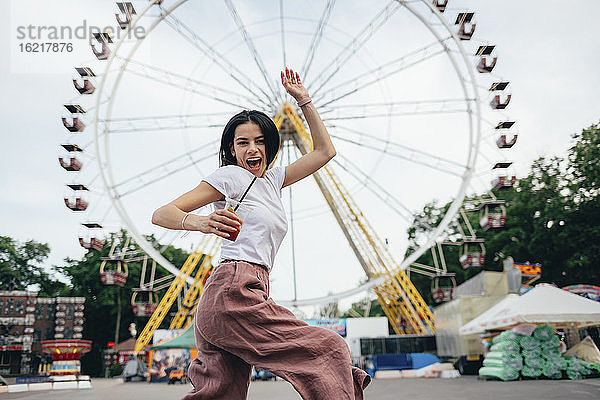 Junge Frau hält Softdrinkbecher und tanzt gegen Riesenrad in Vergnügungspark