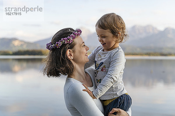 Lächelnde Mutter mit Diadem  die ihre Tochter am Ufer eines Sees trägt