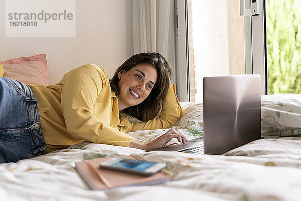 Lächelnde Frau  die einen Laptop benutzt  während sie im Schlafzimmer liegt